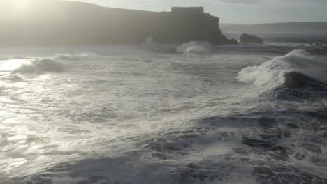 Drone-flyover-powerful-ocean-waves-breaking-toward-Nazare-Lighthouse-promontory,-sunbeam-reflection