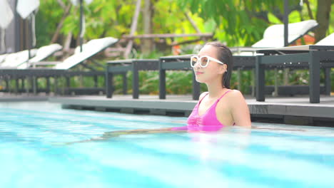 while sitting on a step in the shallow end of a resort pool, a young woman in huge white sunglasses turns her head to look around
