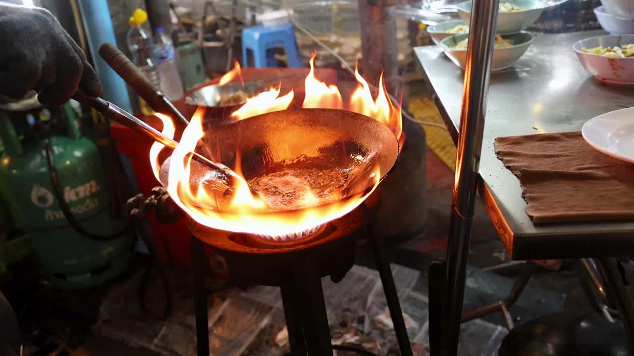 Chef stir-fries ingredients in flaming wok at Bangkok street food stall, dynamic handheld camera movement