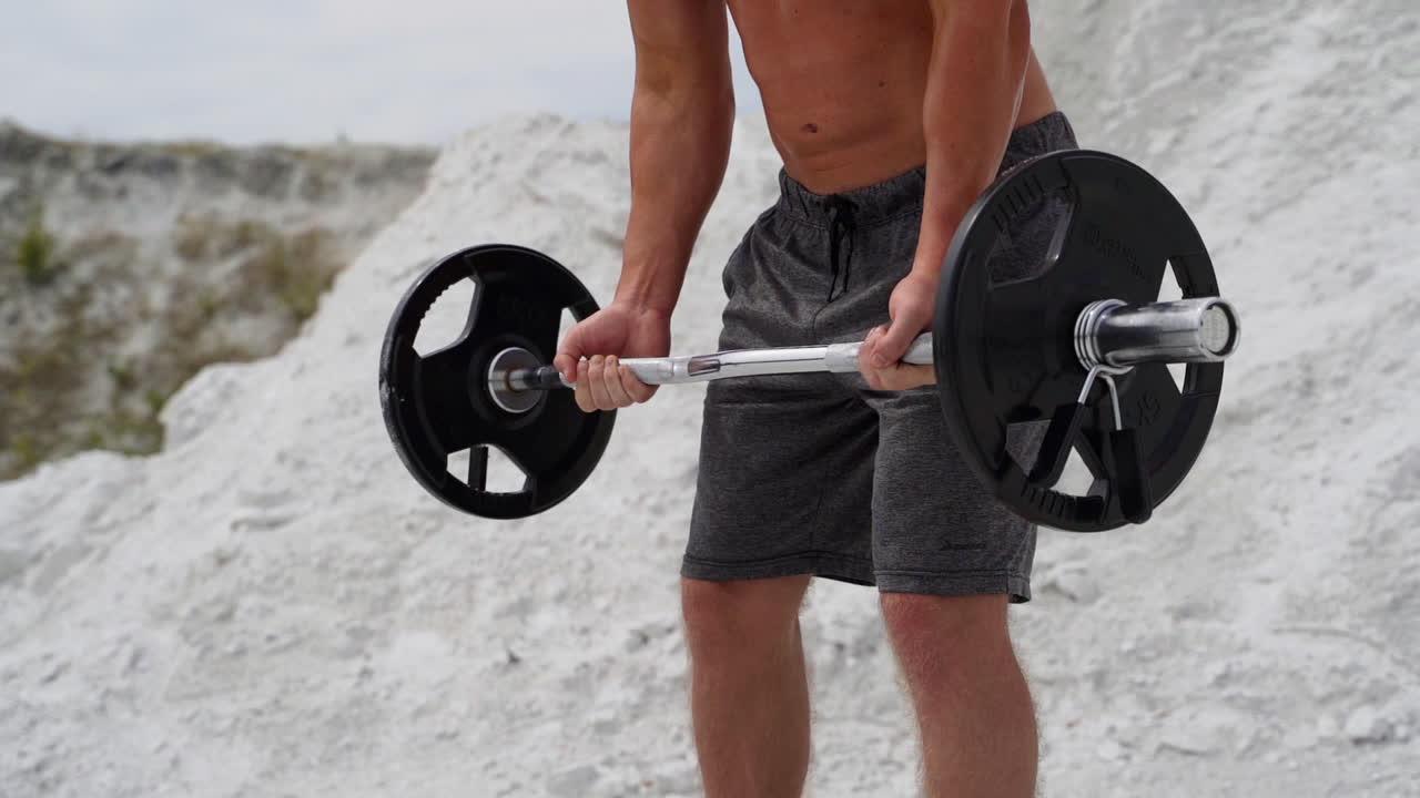 Close-up view of muscular man's body with barbell outdoors. Bodybuilder is doing his workout on the white mountain background.