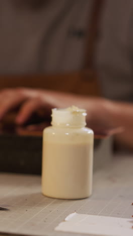 worker applies glue from plastic bottle onto leather detail edges at table closeup. leathersmith works with craft material in handicraft home studio