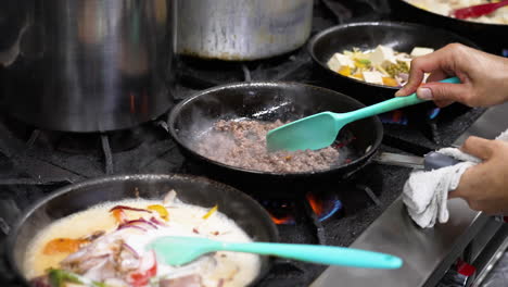 el chef rompe la carne molida en una sartén chisporroteante mientras otras sartenes hierven y humen sobre la llama de la estufa en la cocina de un restaurante comercial, deslizador en cámara lenta de cerca 4k