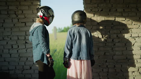 back view two friends wearing helmets stand in an abandoned building, gazing through an opening toward a vast, peaceful field, shadows of their helmets reflect on the brick wall