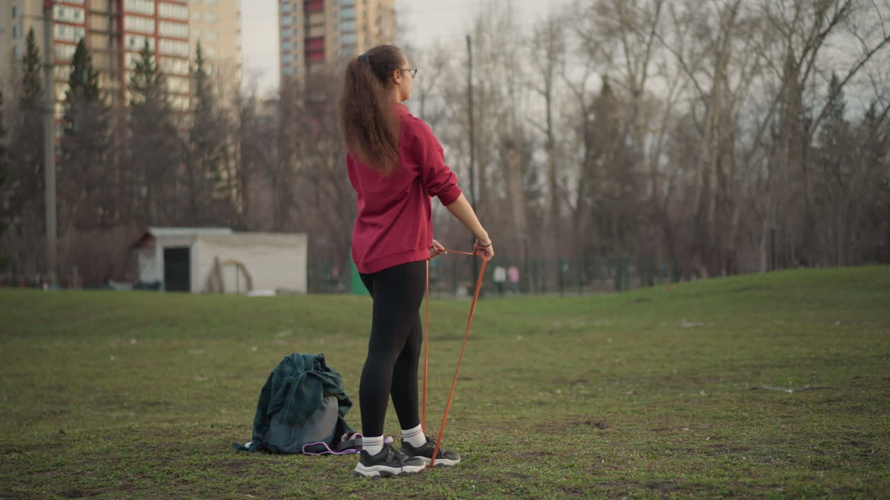 White Woman Exercising With Resistance Band In City Park Near Historic Building, Standing Pulls And Steady Stance, Long Ponytail And Red Hoodie, Urban Trees And Skyline, Solitary Practice With Bag