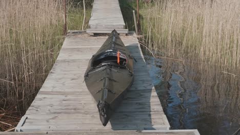 Toma-De-ángulo-Alto-Sobre-Un-Kayak-Negro-Plegable-En-Un-Embarcadero-Cerca-Del-Muelle-Con-Pasto-Seco-Al-Fondo-En-Un-Día-Soleado