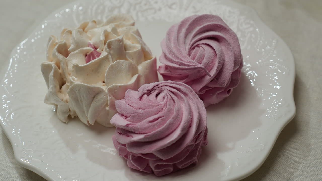 Close up display of decorative white and pink cupcakes arranged on elegant white ceramic plate with delicate patterns, showing swirled icing design on light tablecloth in cozy setting
