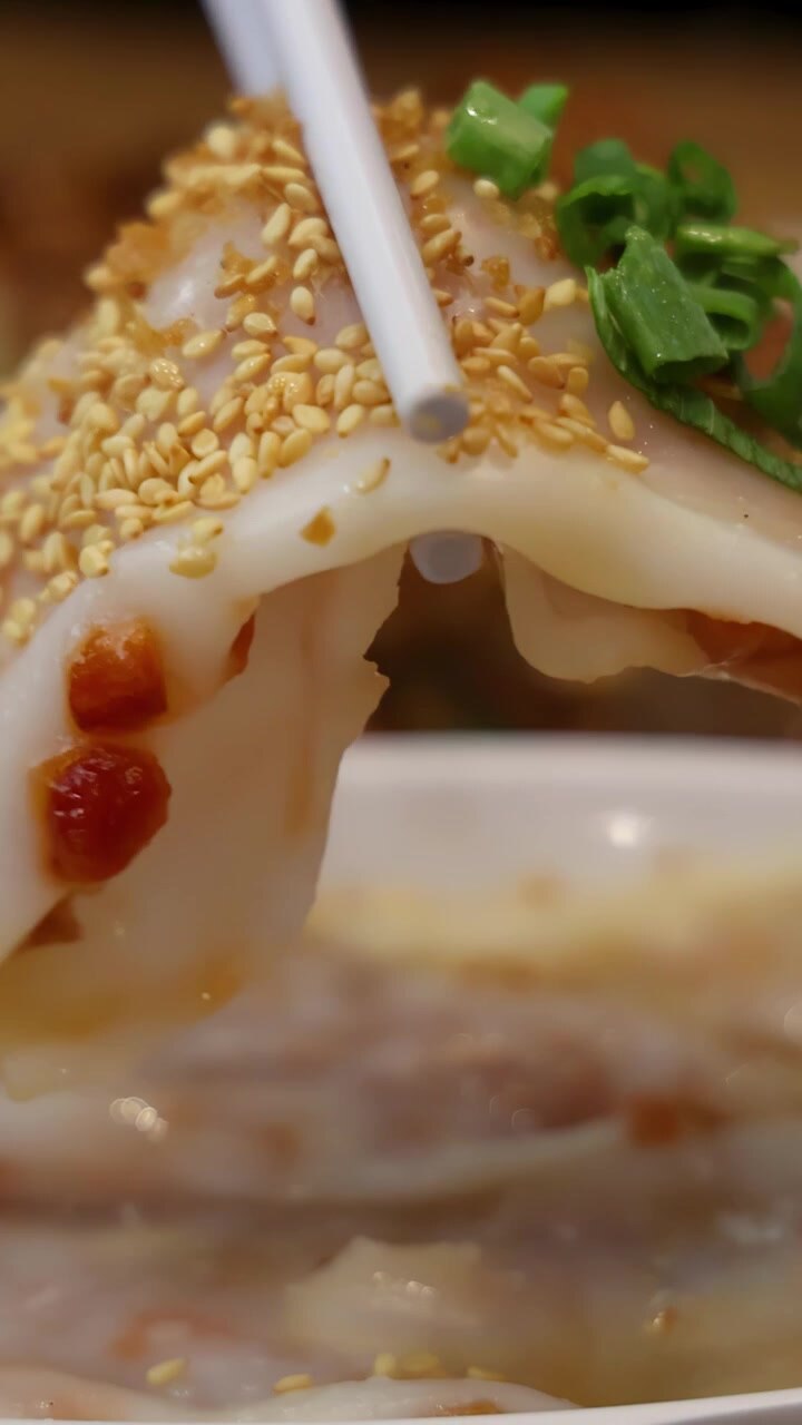 Primo piano di bacchette che raccolgono un involtino di noodle di riso al vapore guarnito con semi di sesamo e cipollotti sotto un'illuminazione calda e naturale in un ambiente da pranzo informale