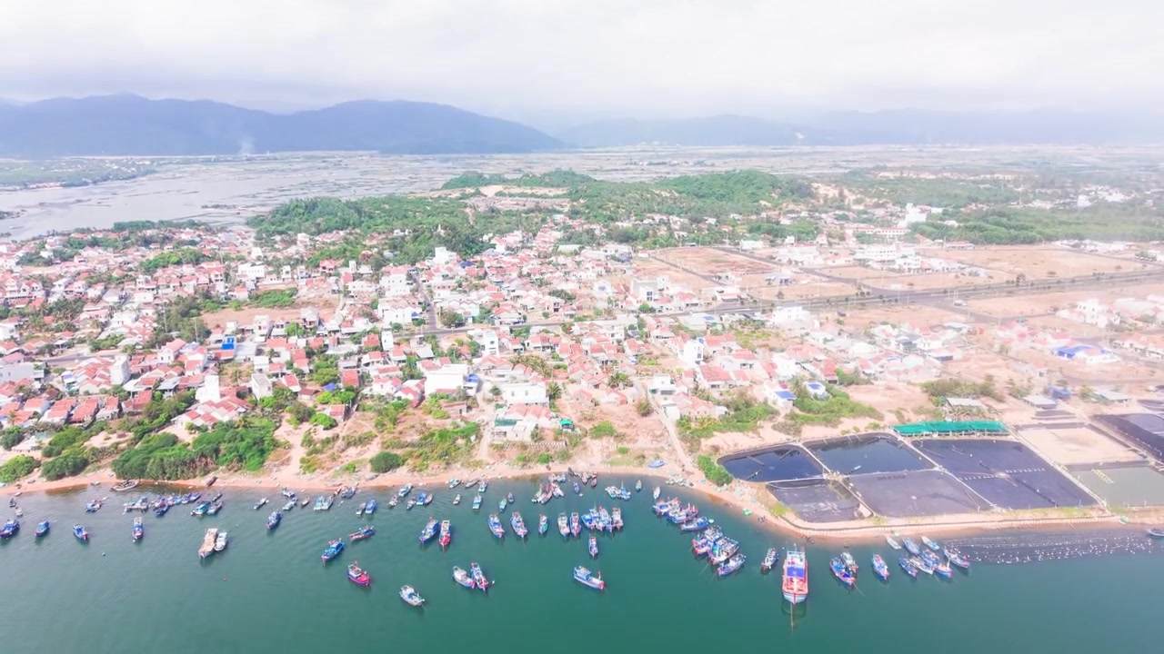 As vistas revelam a paisagem de Bien Lo Quang no Vietnã. A área apresenta uma mistura de fazendas, barcos e pequenas casas à beira da água. O céu está nublado sobre esta comunidade costeira.