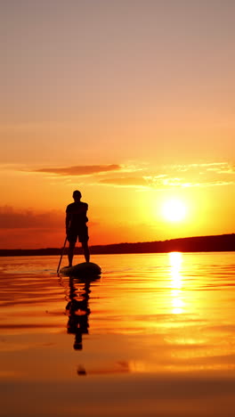 orange sky merging with orange river surface at sunset. male silhouette approaches oaring on the sup board. low angle view. vertical video.