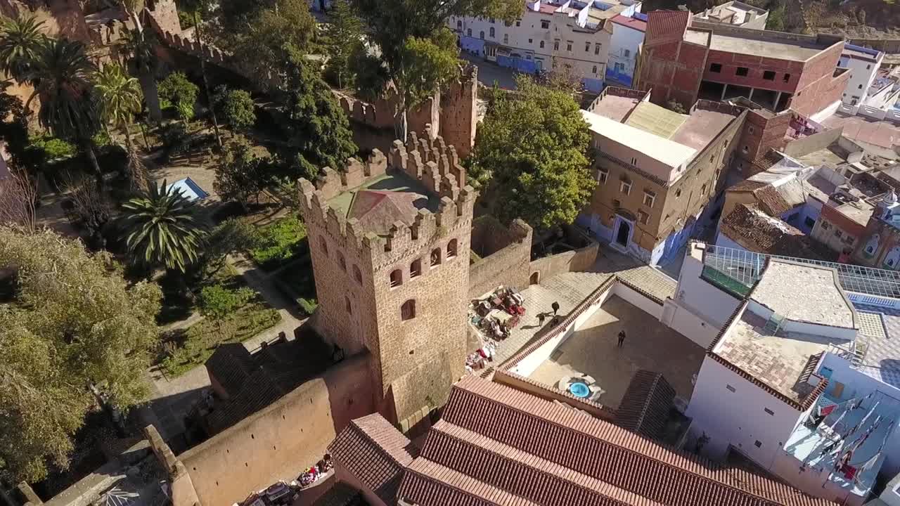 Szczegółowe ujęcie lotnicze HD historycznej wieży Kasbah w Chefchaouen. Podkreśla starożytną architekturę kamienną, bujne zielone ogrody oraz otaczające ją ikoniczne niebieskie budynki medyny.
