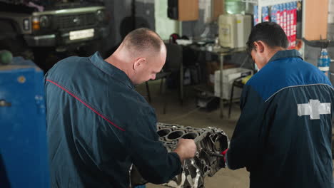 automotive engineers in a workshop, one focused on assembling an engine while his colleague uses a smartphone, amidst mechanical equipment in the background