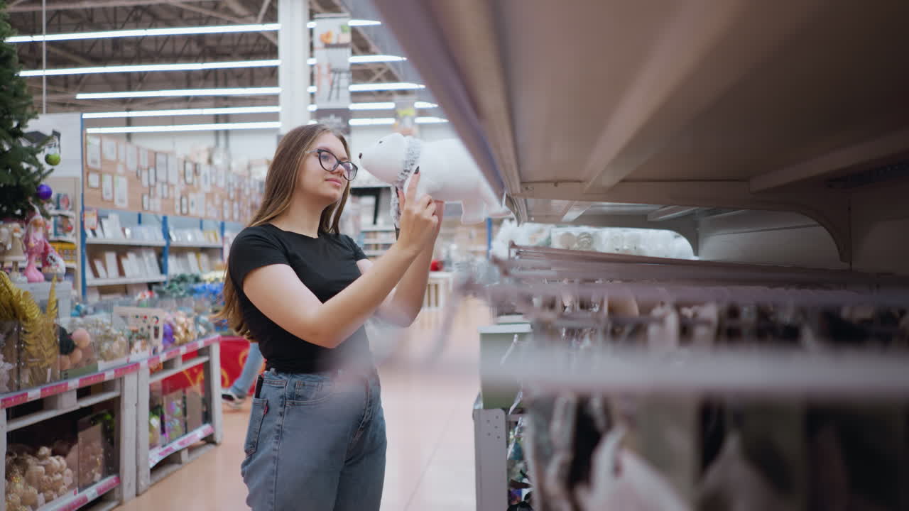 una mujer joven con gafas y camisa negra examina pensativamente un juguete blanco de peluche de oso polar en una tienda de vacaciones, con otro cliente pasando en el fondo en medio de decoraciones festivas