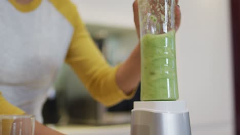 Mixed-race-woman-preparing-healthy-drink,-mixing-fruit-and-vegetables-in-kitchen