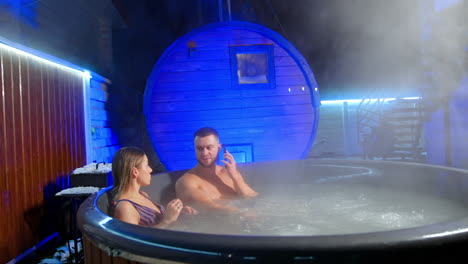 couple spends time together in a spa. caucasian man and woman sit in hot tub relaxing. man uses his phone.