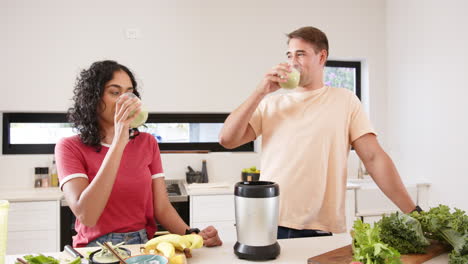 young multiracial couple drinking smoothies in kitchen with blender and fresh fruits, at home