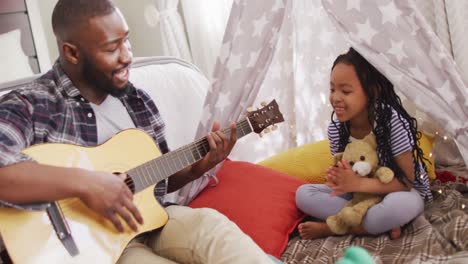 Happy-african-american-father-and-daughter-sitting-in-blanket-tent,-playing-guitar-and-singing