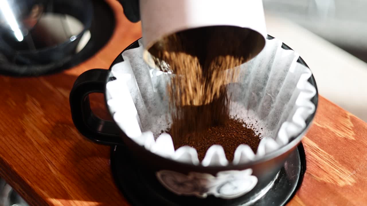 Eine behandschuhte Hand gießt Kaffeepulver in einen Pour-over-Papierfilter bei warmem Licht der goldenen Stunde – statische Nahaufnahme mit reicher Textur, sanftem Bokeh und einem sauberen Arbeitsablauf der Kaffeezubereitung.