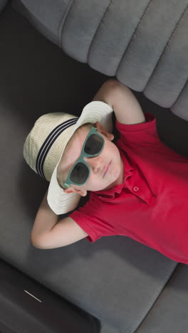 stylish little boy in straw hat enjoys relaxing on couch on yacht. sweet toddler in sunglasses rests in rocking cabin during trip upper close view