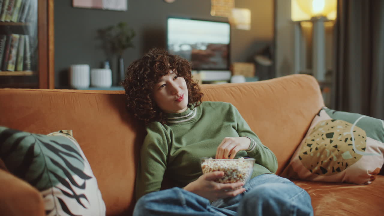 Young Woman Lounging on Sofa, Snacking on Popcorn and Watching Television