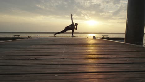 Mujer-Joven-Haciendo-Estiramientos-De-Yoga-En-El-Muelle-Al-Atardecer,-Con-Vistas-Al-Mar-En-Calma