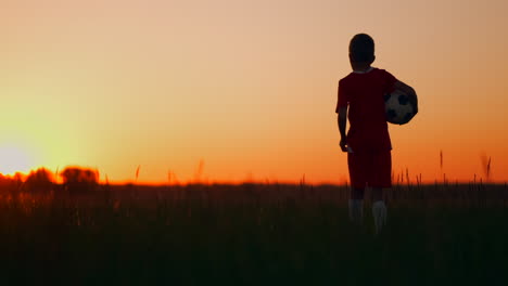 Der-Junge-Geht-Bei-Sonnenuntergang-Mit-Einem-Fußball-Auf-Das-Feld,-Blickt-In-Die-Ferne-Und-Träumt-Davon,-Ein-Erfolgreicher-Fußballspieler-Zu-Werden.-Beobachten-Sie-Den-Sonnenuntergang-Auf-Dem-Feld