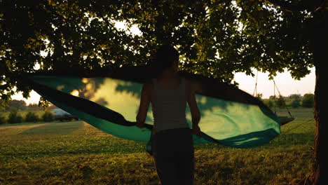 woman shakes out a hammock hanging between two trees as the sun sets in the background