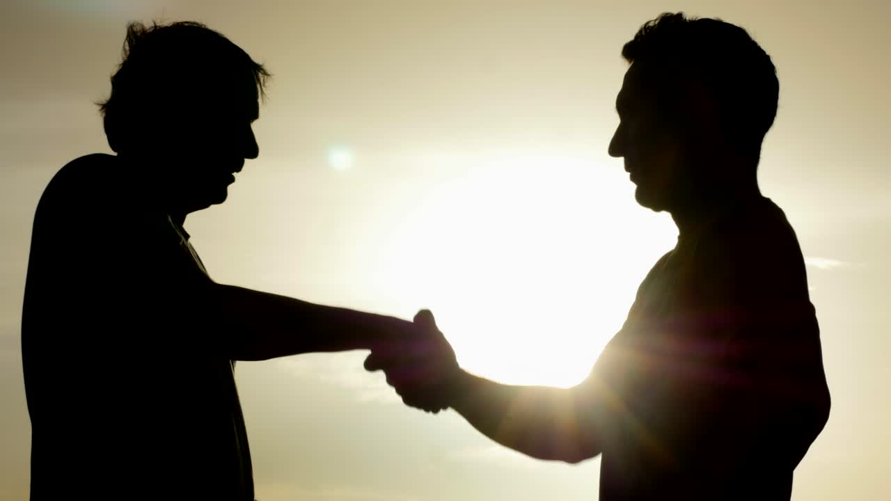 Silhouetted men engage in a humorous handshake against a warm sky backdrop