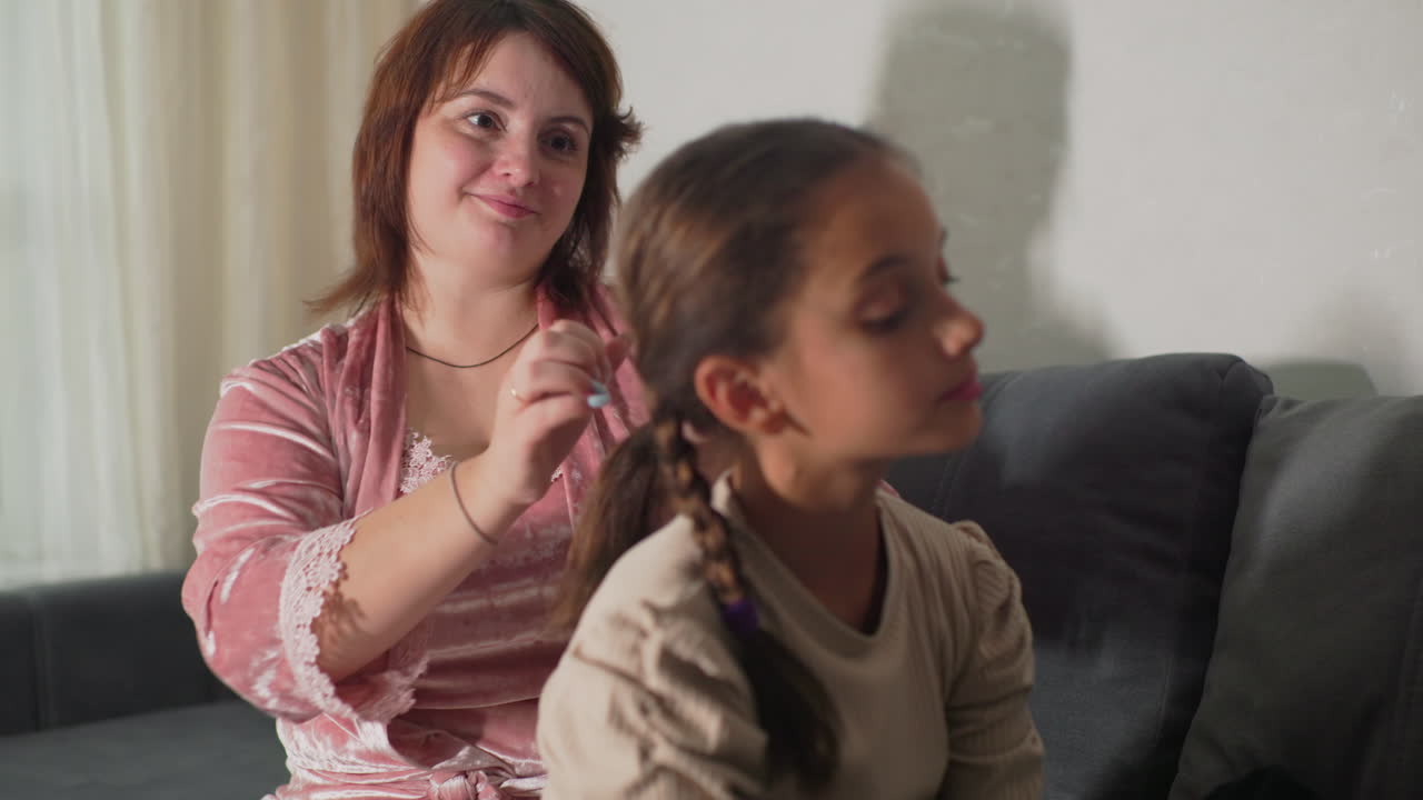 Mum braiding daughter’s hair in cozy living room, close-up of caring hands weaving neat braided pigtail, child relaxed on sofa, soft lighting highlighting tender family bond and intimate personal grooming moment