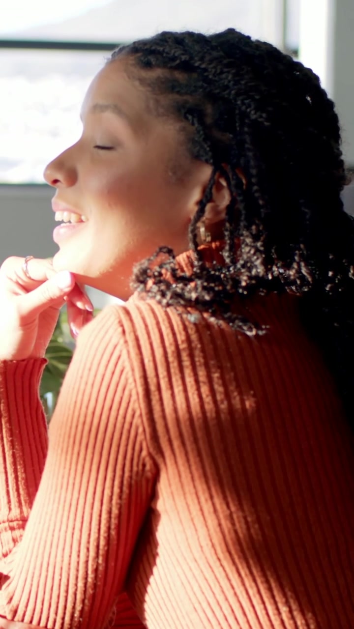 Vertical video: Sunlight on face as woman in orange sweater gazing out window at desk, copy space