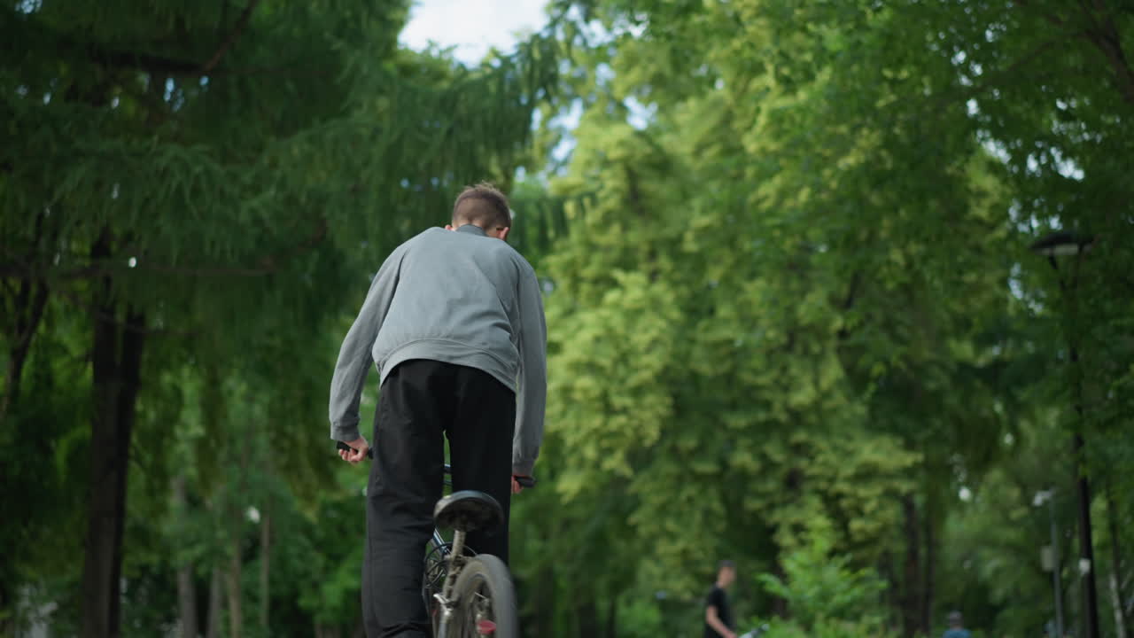 불규칙한 패턴으로 자전거를 타는 동안 서있는 회색 을 입은 소년의 뒷면, 배경에는 울창한 녹색 나무, 누군가 서있는, 그리고 멀리 있는 건물의 보케 효과가 있습니다.