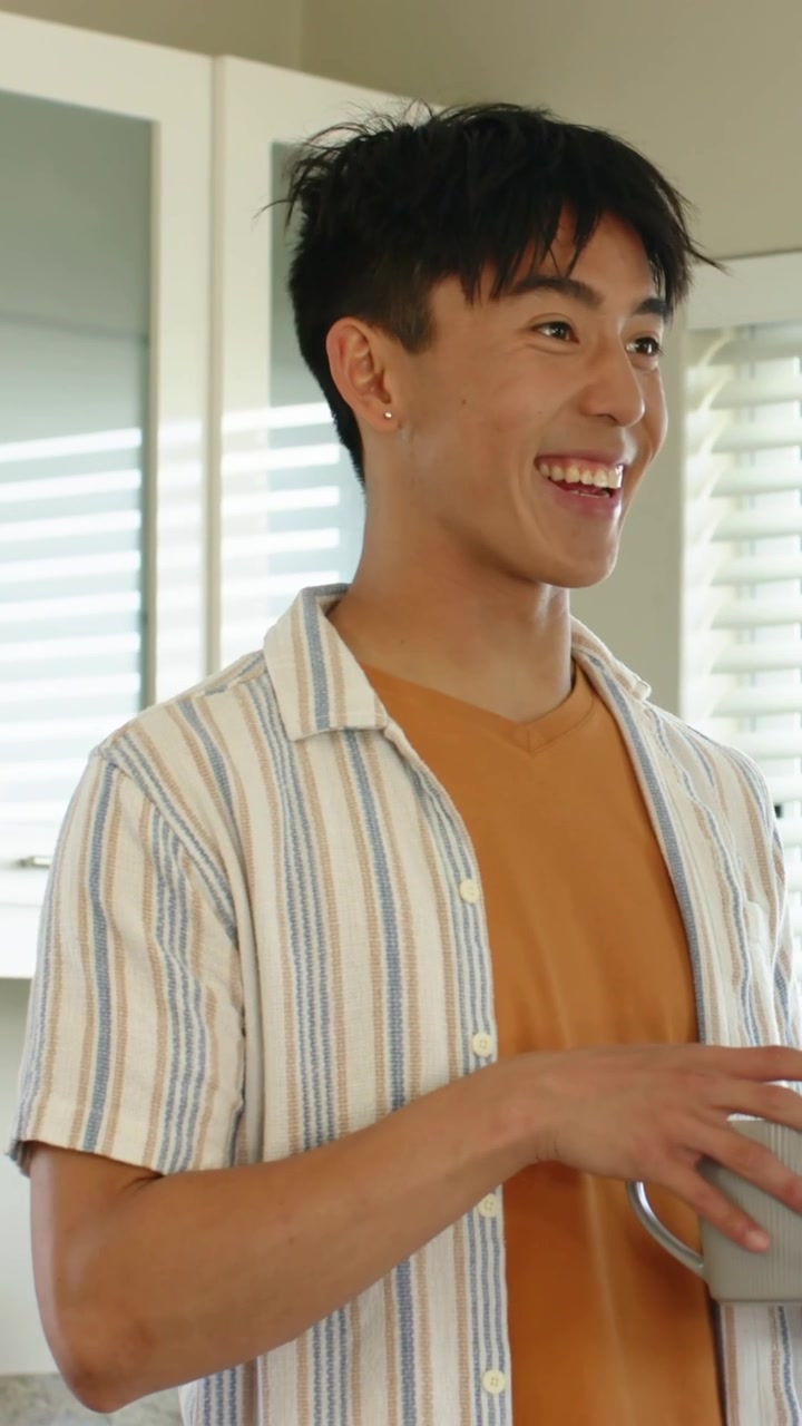 縦型動画：キッチンで、オレンジのTシャツを着た男性がグレーのマグカップを抱え、声に反応してちらりと見て微笑んでいる。