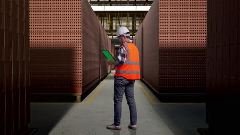 full body back view of asian male engineer with safety helmet working on a green screen tablet and looking around while standing with red brick packed in stacks are stored