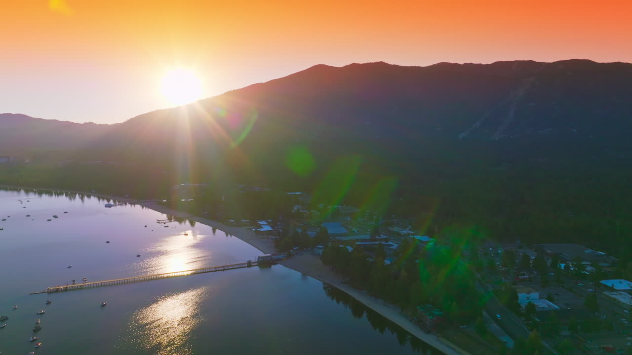 Last rays of setting sun peeping from behind the high mountains. Beautiful lake with urban infrastructure at the shore. Orange sky at backdrop.