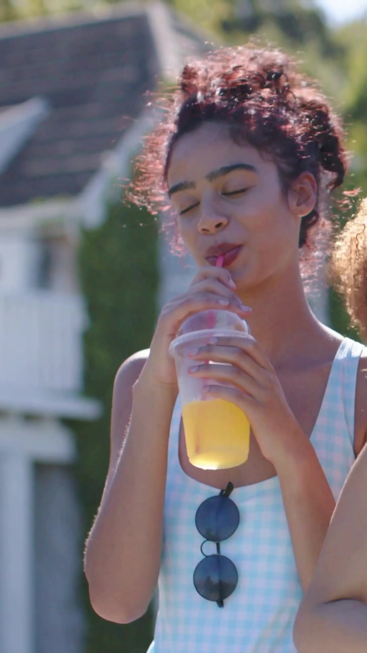 Video vertical: Mujer con traje azul levantando una taza, bebiendo en el porche, refrescándose con pajita rosa y gafas de sol.