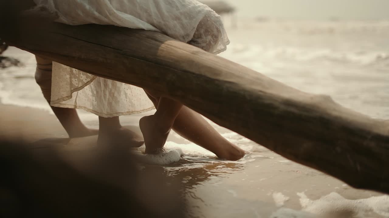 Closeup shot of a waves hitting legs of a couple sitting beside seashore on beach.