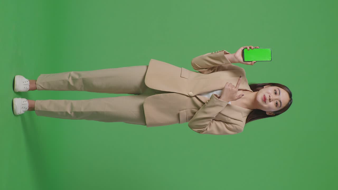 полное тело азиатской бизнесменки с улыбкой, показывающей и указывающей на зеленый экран мобильного телефона, стоя перед зеленым экраном