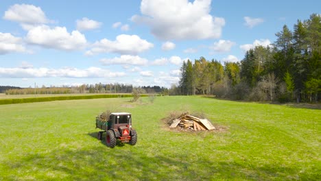 aerial drone footage flying behind and old retro vintage tractor that is driving with garden waste or tree branches in its box during a summer sunny day in a green grassy countryside farmland.