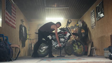 A-male-mechanic-with-a-short-haircut-in-a-gray-T-shirt-removes-his-bike-from-the-jack-and-takes-it-out-of-his-garage-workshop.-Male-mechanic-completing-motorcycle-repair