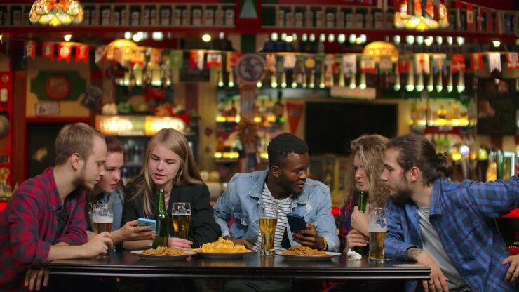 A man shows his friends a photograph on the phone and everyone laughs while paired and drinking beer.
