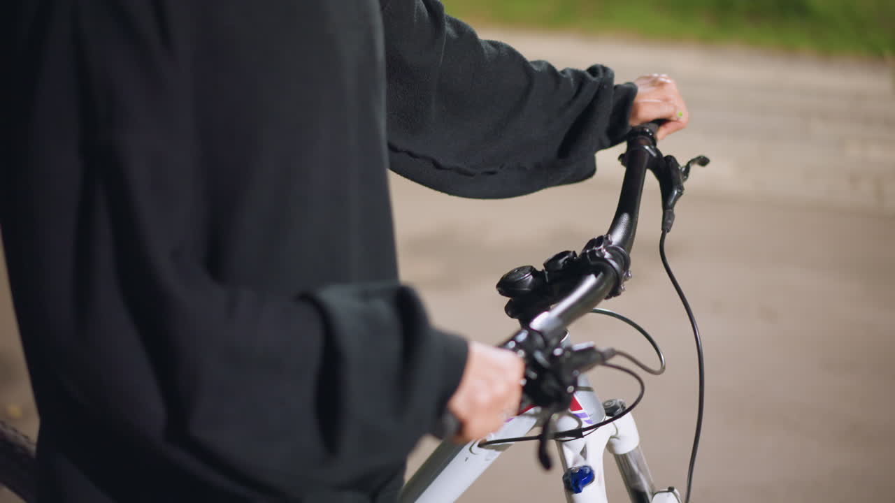 Pavement CloseUp Of Cyclist Holding Handlebars At Night, Person In Loose Black Hoodie Stands Near Curb With Hands On Grips, Ambient Lamplight And Soft Background, Stillness And Reflective Mood