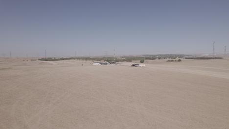 Tiendas-De-Campaña-Beduinas-En-Un-área-árida-Y-Remota,-En-Un-Campo-De-Arena-Seco-Fuera-De-La-Red,-Cerca-De-Grandes-Líneas-Eléctricas-Y-Una-Fábrica-De-Alimentos-Para-Animales:-Toma-De-Paralaje