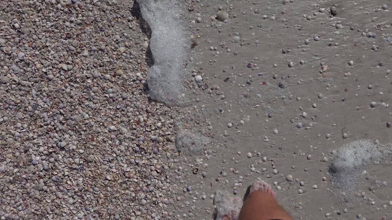 A barefoot person steps forward across textured wet sand scattered with small shells as white sea foam rolls over the shoreline. The scene shows coastal detail.