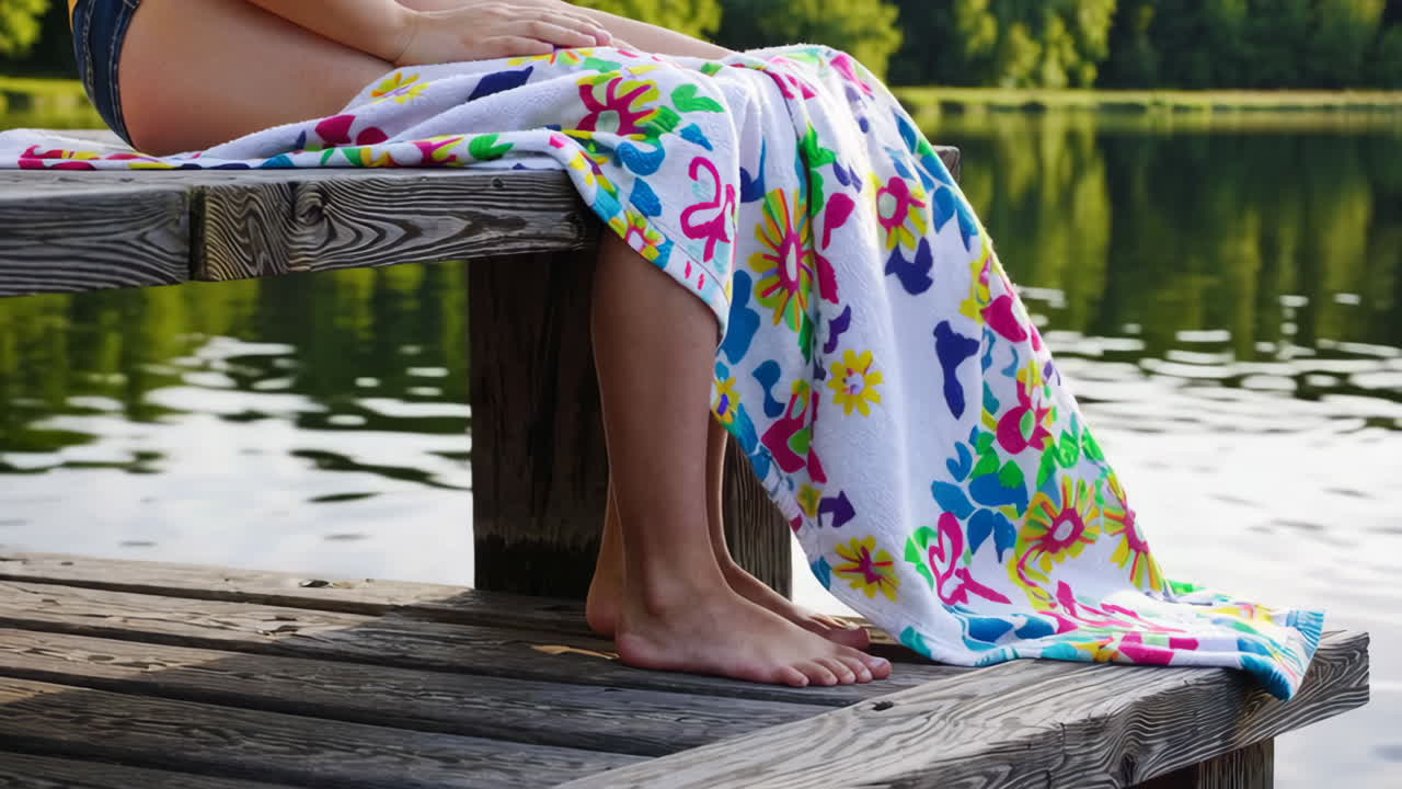 Person wrapped in a colorful towel sitting on a wooden dock by a lake