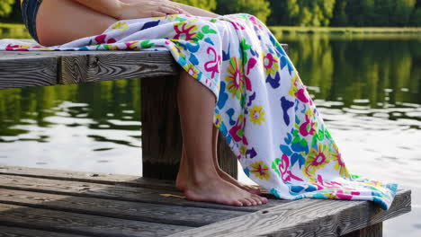 person wrapped in a colorful towel sitting on a wooden dock by a lake