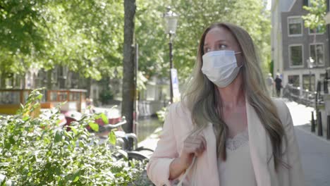 Attractive-Blond-Woman-With-Mask-Walking-On-Stone-Pavement-In-Amsterdam-Canal,-Netherlands-During-Covid-19-Pandemic