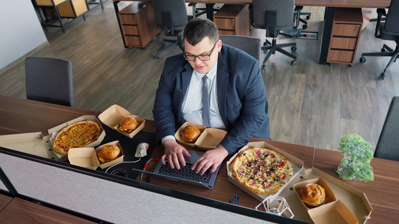 Mand i jakkesæt skriver på bærbar computer omgivet af fastfood på kontoret