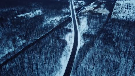 Toma-Aérea-De-Un-Desolado-Camino-Invernal-Frío-En-Un-Bosque-Oscuro-Con-Vista-Al-Río