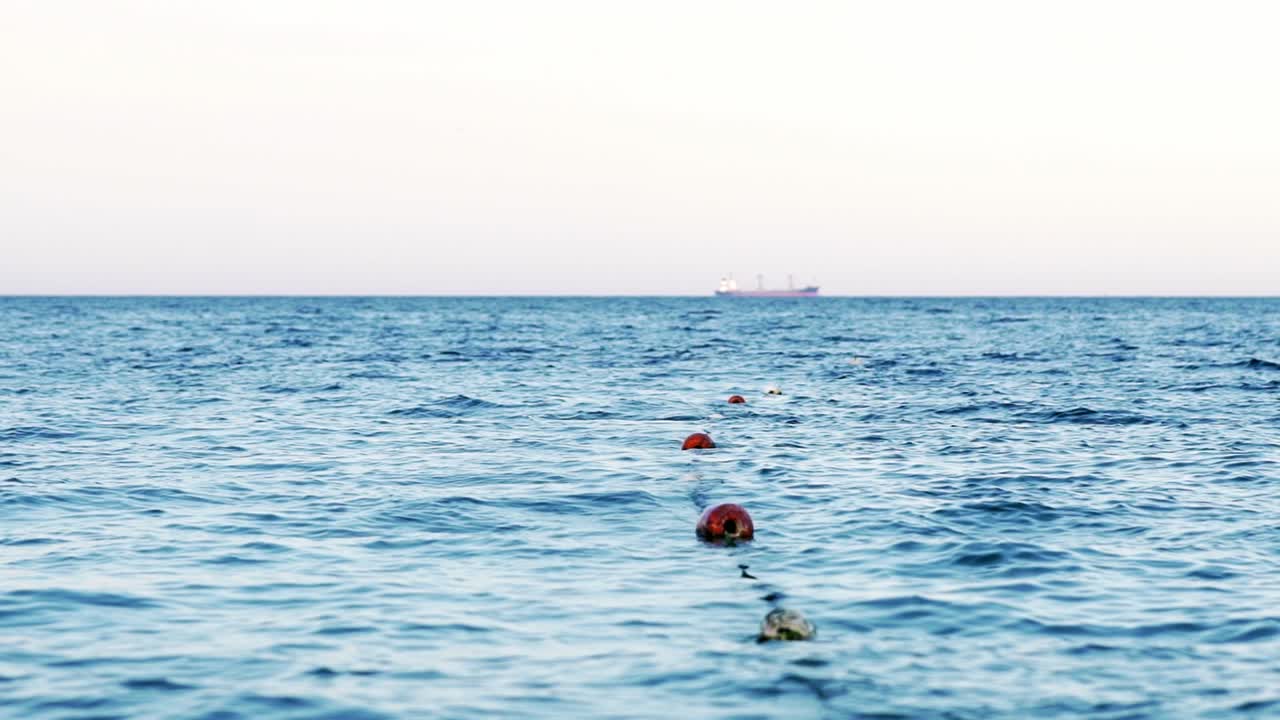 Amazing view of blue sea water with buoys on the rope. Safe swimming zone with markers on the sea. Big ship floating beyond afar. Slow motion.