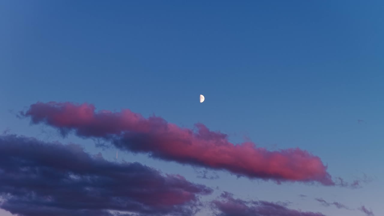 First quarter moon against a blue sky with cloud streaks glowing in soft sunset hues