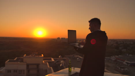 Un-Hombre-Con-Audífonos-En-El-Techo-Se-Relaja-Trabajando-Remotamente-Disfrutando-De-La-Vida-A-Pesar-De-Tomar-Una-Cerveza-Y-Escribir-En-El-Teclado.-Opere-En-La-Bolsa-De-Valores-Usando-Una-Computadora-Portátil-Y-Disfrutando-De-La-Hermosa-Vista